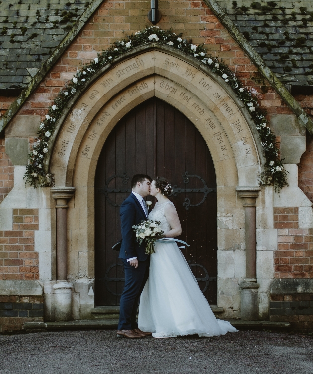Couple kiss underneath church arch dressed with flowers