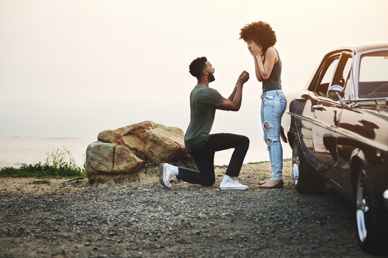 man kneeling on one knee on cliff top proposing to girlfriend next to car